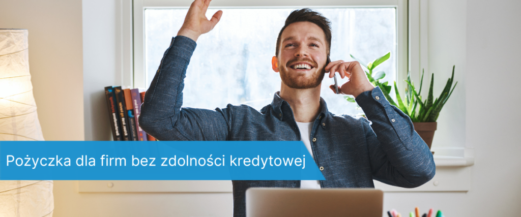 Uśmiechnięty mężczyzna siedzący przy biurku z laptopem, wykonujący gest triumfu podczas rozmowy telefonicznej